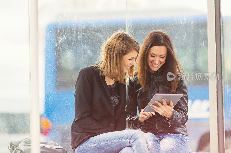 Two friends using digital tablet on bus station